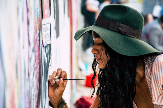 A woman wearing a hat paints a colourful wall with a fine paitnbrush